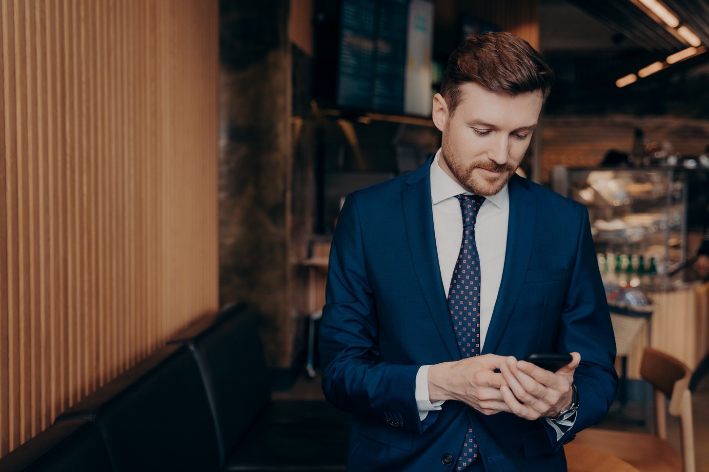 Focused male investor in blue suit being notified about stock market prices, checking his phone, looking at messages and deciding on if to invest or not, standing alone in cafe